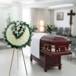 Ataúd de madera barnizada con detalles metálicos y paño blanco, junto a una corona fúnebre redonda de flores blancas sobre caballete en una sala velatoria iluminada