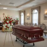 Ataúd de madera oscura sobre carro metálico junto a arreglo floral de rosas rojas y blancas en una sala de velatorio