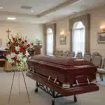 Ataúd de madera oscura sobre carro metálico junto a arreglo floral de rosas rojas y blancas en una sala de velatorio