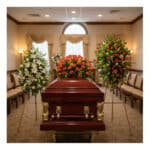 Ataúd de madera barnizada con detalles dorados rodeado de tres grandes arreglos florales en una sala de velación
