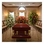 Ataúd de madera barnizada con detalles dorados rodeado de tres grandes arreglos florales en una sala de velación