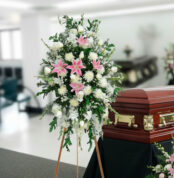 Arreglo floral fúnebre alto con lirios rosas y flores blancas junto a un ataúd de madera en una sala de velación
