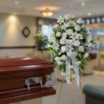 Arreglo floral fúnebre de flores blancas sobre caballete al lado de un ataúd de madera barnizada en una sala de velatorio