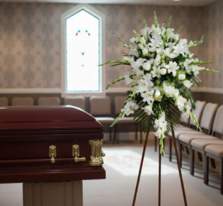Ataúd de madera barnizada junto a corona de flores blancas en una sala de velatorio con vitrales y filas de sillas