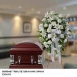 Arreglo floral funerario de pie con flores blancas y lazos, colocado junto a un ataúd de madera oscura en una sala velatoria iluminada