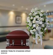 Arreglo floral funerario de pie con flores blancas y lazos, colocado junto a un ataúd de madera oscura en una sala velatoria iluminada