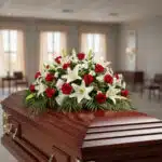 Centro de flores fúnebres con rosas rojas y lirios blancos colocado sobre un ataúd de madera barnizada en una sala velatoria