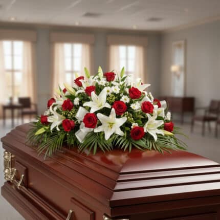 Centro de flores fúnebres con rosas rojas y lirios blancos colocado sobre un ataúd de madera barnizada en una sala velatoria
