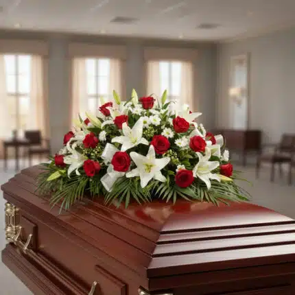 Centro de flores fúnebres con rosas rojas y lirios blancos colocado sobre un ataúd de madera barnizada en una sala velatoria