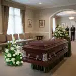 Ataúd de madera oscura con arreglos florales blancos en una sala de velatorio elegante