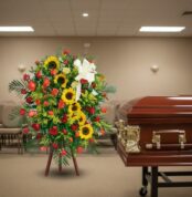 Arreglo fúnebre vertical con girasoles amarillos y rosas rojas sobre caballete, colocado al lado de un ataúd de madera barnizada en una sala velatoria