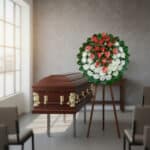 Ataúd de madera marrón con detalles dorados junto a corona fúnebre de flores blancas y rojas sobre caballete en una sala de velatorio iluminada por una gran ventana