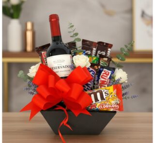 Arreglo floral AMADEA con rosas blancas, eucalipto, lavanda, vino tinto, chocolates variados y lazo rojo en cesta negra.