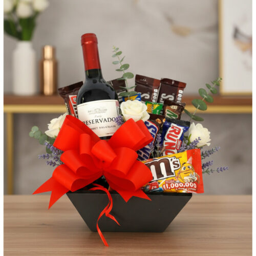 Arreglo floral AMADEA con rosas blancas, eucalipto, lavanda, vino tinto, chocolates variados y lazo rojo en cesta negra.