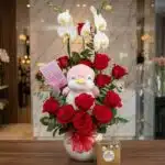 Arreglo floral ALICE con orquídeas blancas, rosas rojas vibrantes, un peluche de pingüino rosa y chocolates Ferrero Rocher, s
