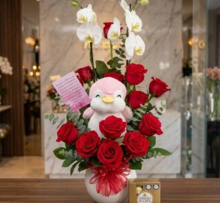 Arreglo floral ALICE con orquídeas blancas, rosas rojas vibrantes, un peluche de pingüino rosa y chocolates Ferrero Rocher, s