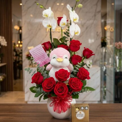 Arreglo floral ALICE con orquídeas blancas, rosas rojas vibrantes, un peluche de pingüino rosa y chocolates Ferrero Rocher, s