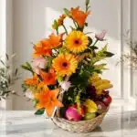 Arreglo Basilio, cesta frutal con rosas, lirios naranjas, gerberas amarillas, uvas, plátanos, manzanas, kiwi y pitahaya sobre