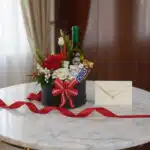 Arreglo floral CARLO con rosas rojas y blancas, vino, chocolates, tarjeta de regalo y cinta roja sobre mesa de mármol.