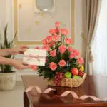Manos delicadas colocando una tarjeta con lazo rosa en una elegante cesta de rosas salmón y frutas frescas sobre una mesa de