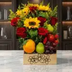 Arreglo floral Adriano con rosas rojas, girasoles vibrantes, naranjas, peras, uvas y fresas en una caja de madera sobre mármo