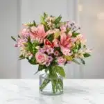 Arreglo floral ANNA con rosas y lirios rosados, alstroemerias y margaritas, en un jarrón de cristal sobre una mesa de mármol