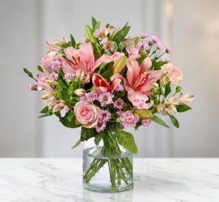Arreglo floral ANNA con rosas y lirios rosados, alstroemerias y margaritas, en un jarrón de cristal sobre una mesa de mármol