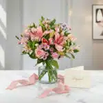 Arreglo floral ANNA con rosas rosadas, lirios y alstroemerias en jarrón de cristal, sobre mármol blanco con tarjeta de regalo