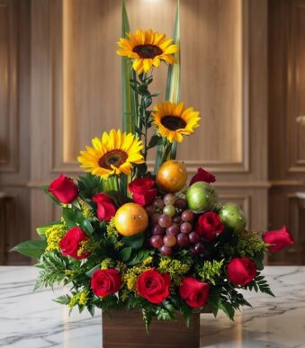 Arreglo floral Batista con rosas rojas, girasoles vibrantes, uvas, naranjas y manzanas verdes, sobre una mesa de mármol en un