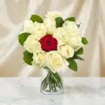 Arreglo floral CARLA con 12 rosas de tallo largo, una roja central y once blancas, en un jarrón de cristal sobre mármol.
