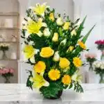 Arreglo floral exótico Amaretto con lirios y rosas amarillas vibrantes, gerberas y follaje verde, sobre una superficie de már