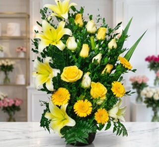 Arreglo floral exótico Amaretto con lirios y rosas amarillas vibrantes, gerberas y follaje verde, sobre una superficie de már