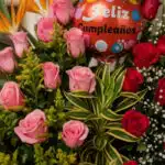 Primer plano extremo de un arreglo floral exótico con rosas rosadas y rojas, follaje verde y un globo rojo de 'Feliz Cumpleañ
