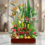 Arreglo floral Akebia con aves del paraíso, rosas rojas, lirios, orquídeas y frutas frescas sobre mármol, fondo de floristerí