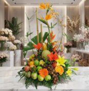 Arreglo floral con frutas exóticas, lirios, aves del paraíso y gerberas en tonos naranjas y amarillos, sobre mármol blanco en