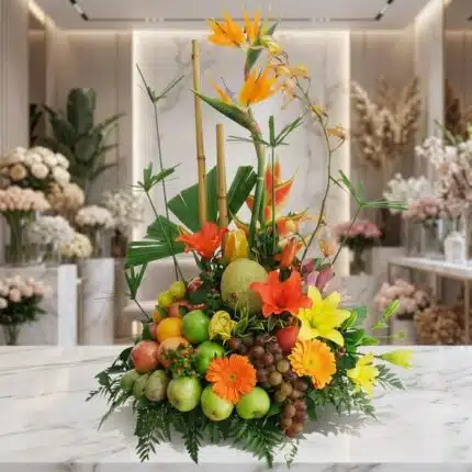 Arreglo floral con frutas exóticas, lirios, aves del paraíso y gerberas en tonos naranjas y amarillos, sobre mármol blanco en