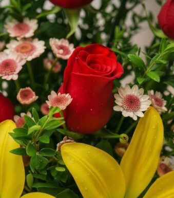 Primer plano extremo de un arreglo floral Capella con una rosa roja central, lirios amarillos y pequeñas margaritas, mostrand