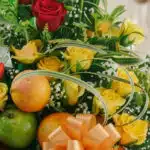 Primer plano extremo de un arreglo floral con rosas amarillas y rojas, manzanas verdes y naranjas, con sutiles gotas de rocío