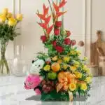 Arreglo Floral con Frutas Encanto, con rosas rojas y amarillas, aves del paraíso, uvas, manzanas, naranjas y un peluche, sobr