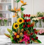 Arreglo floral con frutas y vino, girasoles, rosas rojas y aves del paraíso sobre mármol, estilo estudio premium.