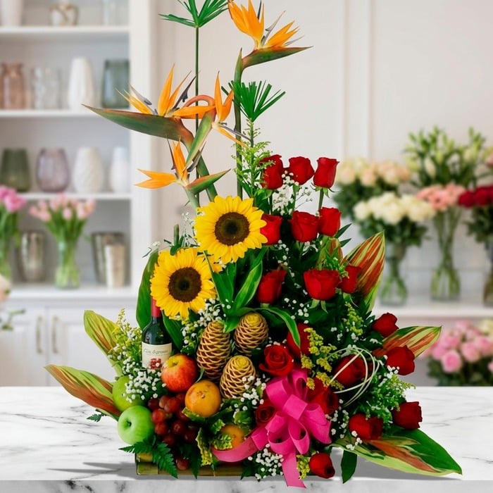 Arreglo floral con frutas y vino, girasoles, rosas rojas y aves del paraíso sobre mármol, estilo estudio premium.