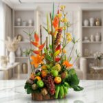 Arreglo floral con frutas exóticas, lirios naranjas, orquídeas amarillas y heliconias rojas, sobre base de madera rústica en