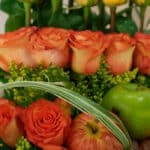 Primer plano extremo de un arreglo floral con rosas naranjas, manzanas verdes y rojas, y hojas verdes, destacando texturas y