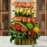 Arreglo floral premium con rosas naranjas y amarillas, manzanas, uvas y naranja, sobre una base de bambú en una superficie de