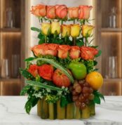 Arreglo floral premium con rosas naranjas y amarillas, manzanas, uvas y naranja, sobre una base de bambú en una superficie de