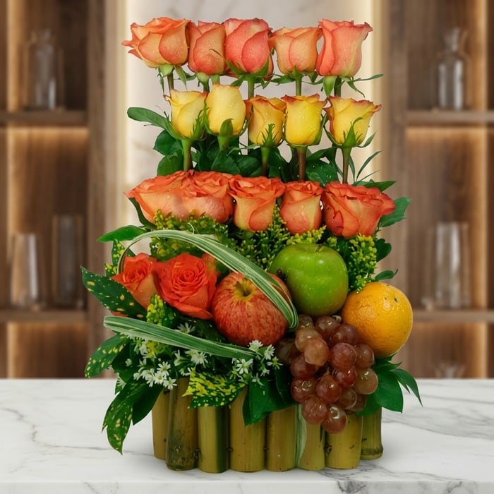 Arreglo floral premium con rosas naranjas y amarillas, manzanas, uvas y naranja, sobre una base de bambú en una superficie de