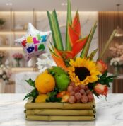 Arreglo floral frutero con tamarillos, uvas, naranjas, girasoles, rosas y heliconias, sobre mármol en floristería premium.