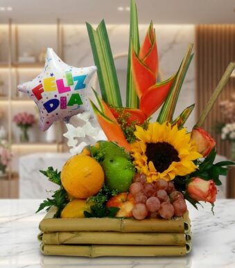 Arreglo floral frutero con tamarillos, uvas, naranjas, girasoles, rosas y heliconias, sobre mármol en floristería premium.