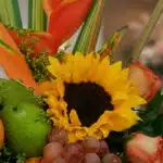 Primer plano extremo de un arreglo floral con girasol, uvas, manzana verde y flores exóticas, destacando texturas y gotas de