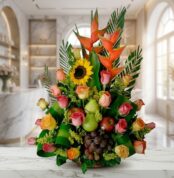 Arreglo floral con rosas de colores, girasoles, heliconias, uvas, peras y manzanas, sobre una superficie de mármol en un ento